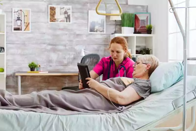 Nurse assisting a bedridden elderly woman using a tablet in a cozy home setting, showcasing home care services in Dunn Loring, VA.